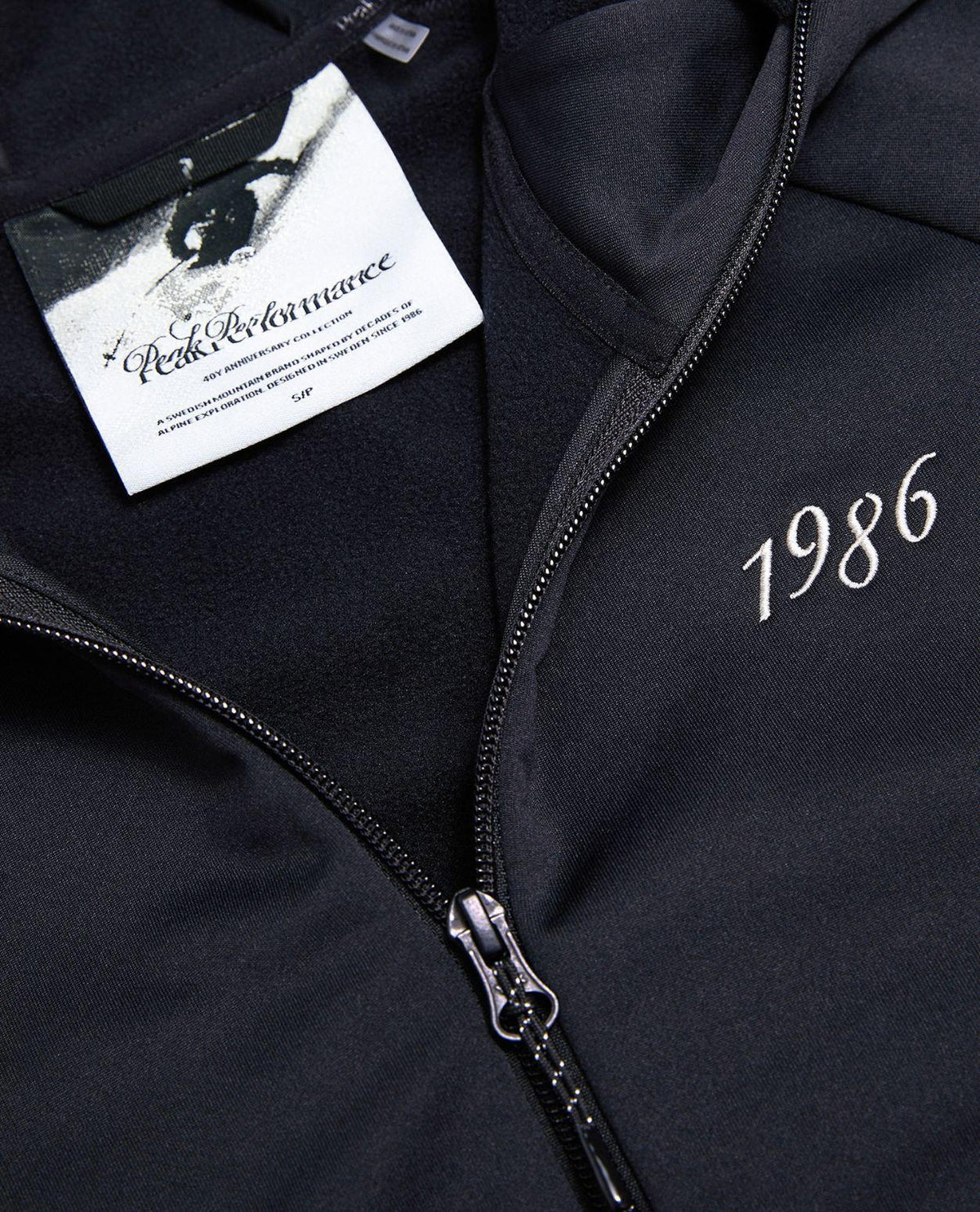Närbild på den svarta Peak Performance 40Y Rider Zip Hood dam med dragkedja och broderat årtal.