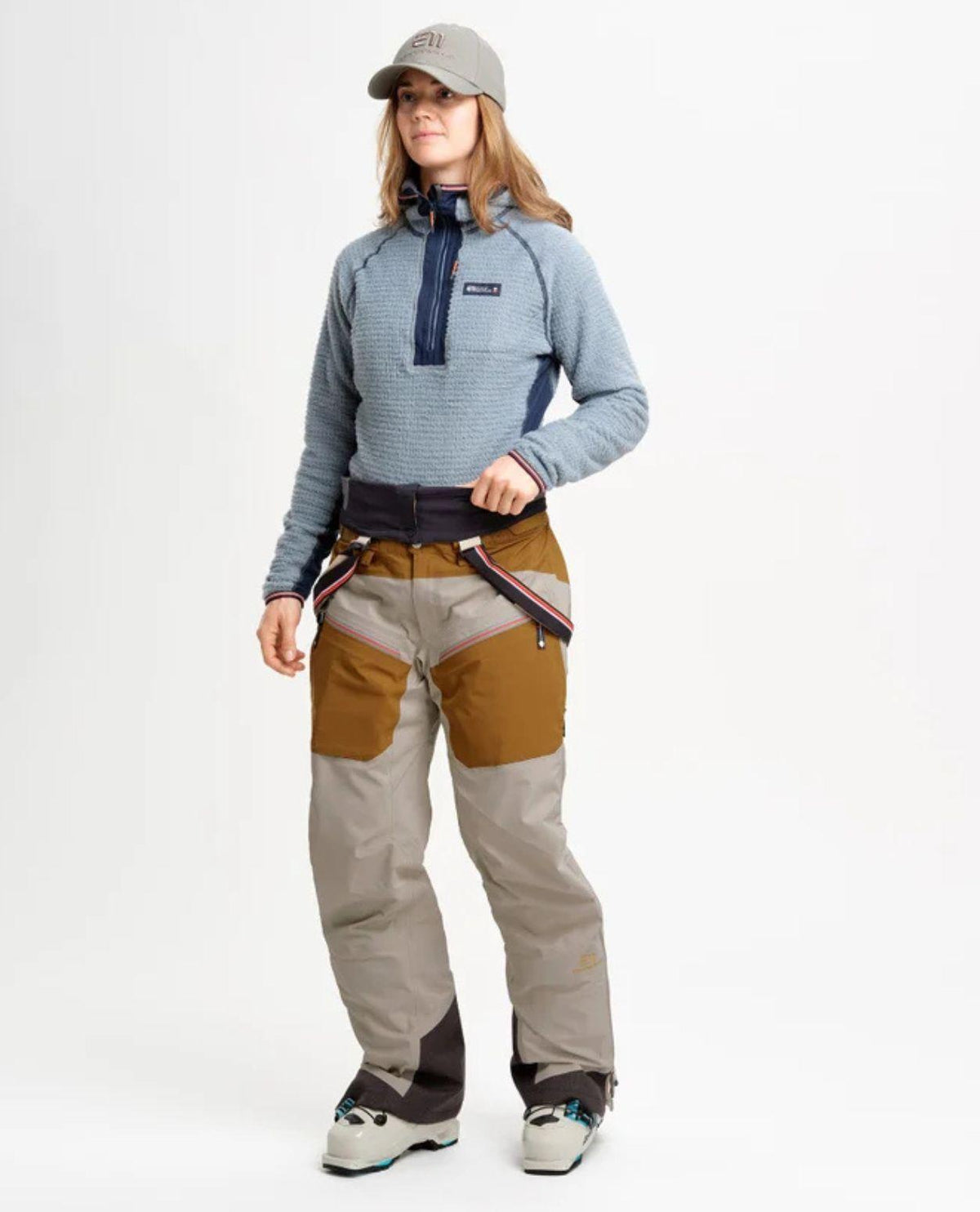 Kvinna som bär en blå mellanlager-tröja, grå keps, brunt och grått skidset samt pjäxor.