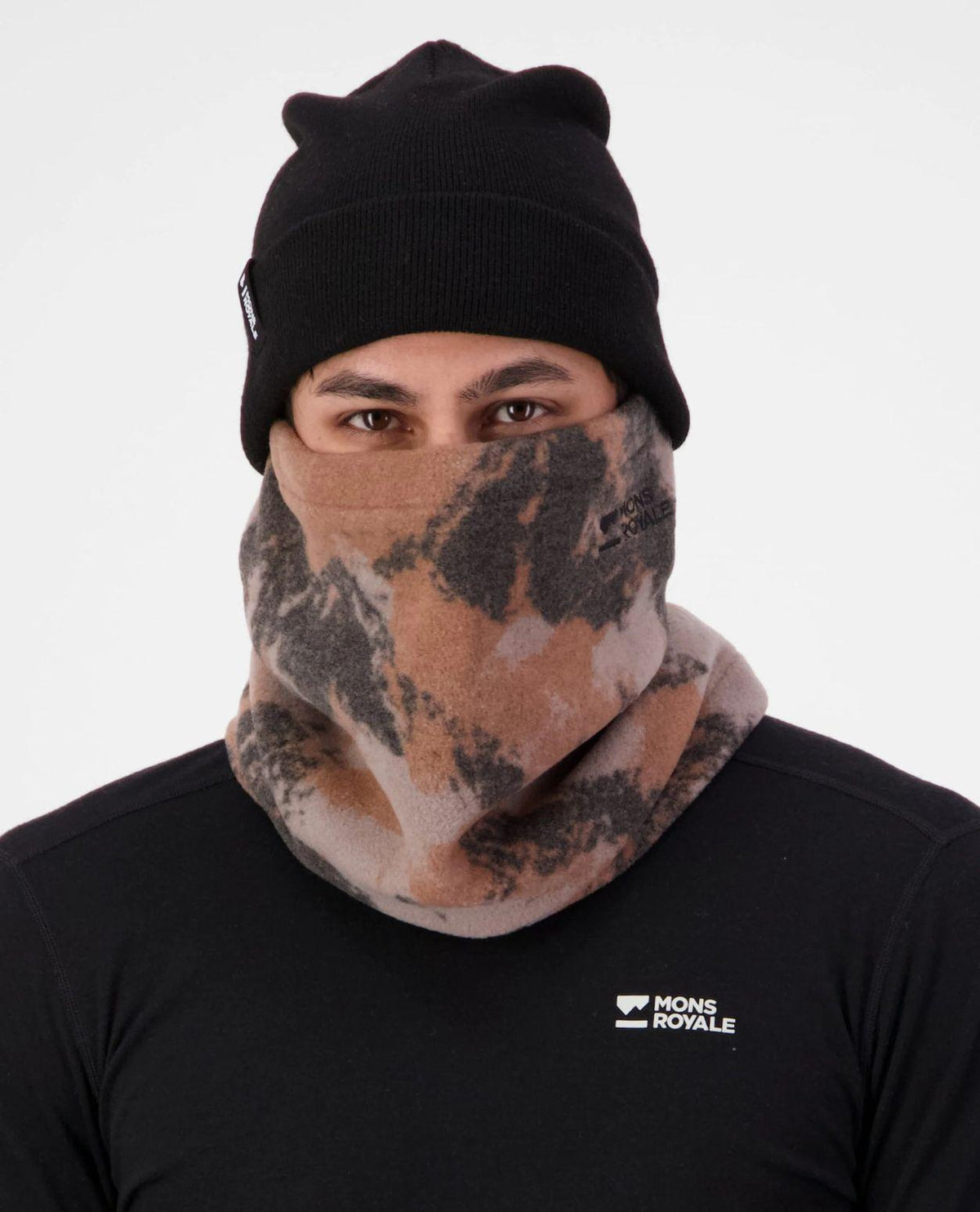 Person bär Mons Royale Pileup Fleece Neck i Mtn Camo Orange samt en svart mössa och tröja.