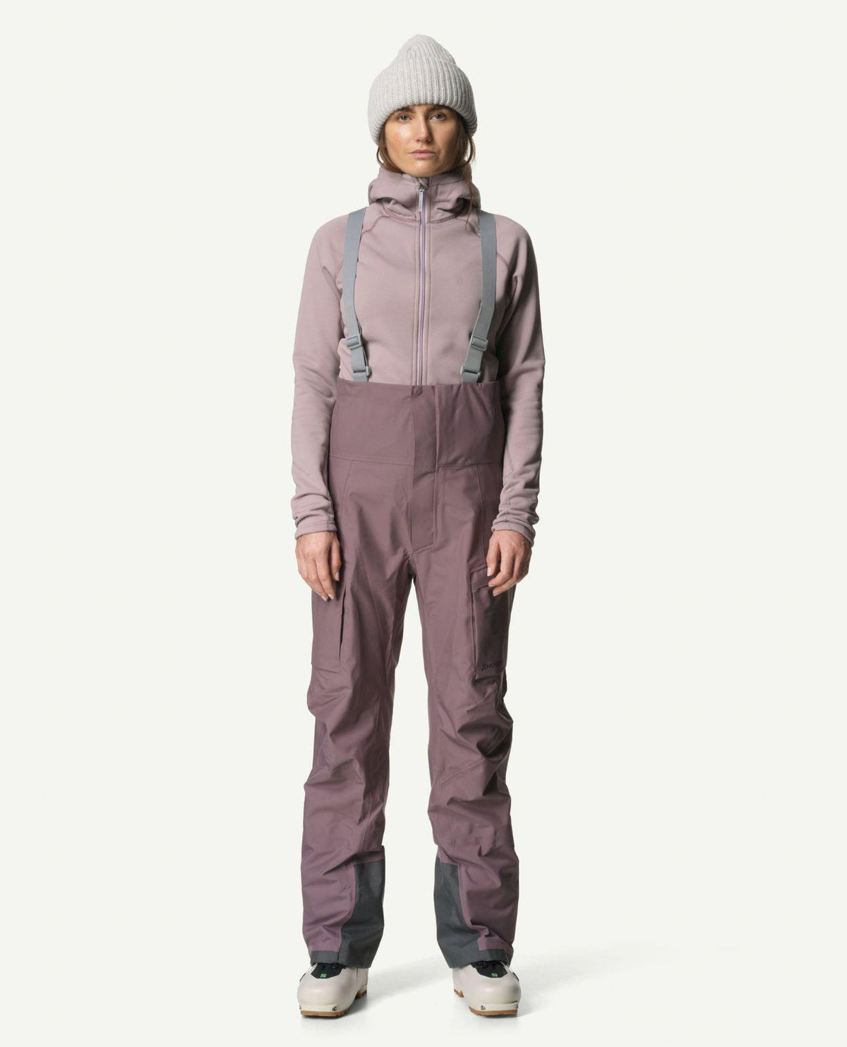 En kvinnlig modell iförd en lila Houdini Power Up Houdi mellanlagringströja, matchande hängselbyxor och en ljus mössa.