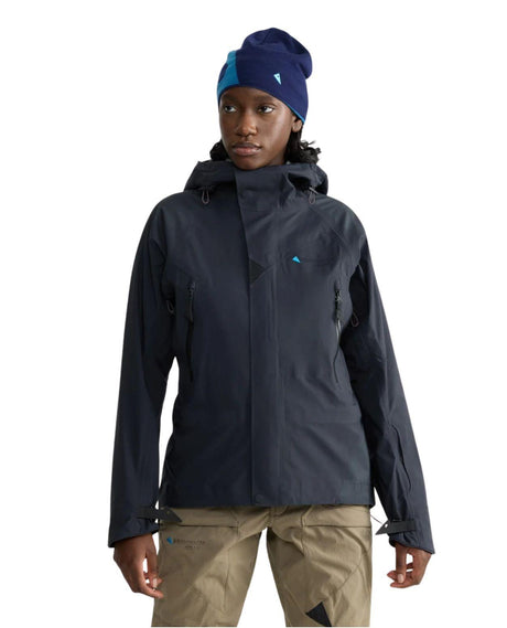 En modell i Klättermusen Svalinn Hood Jacket för dam i svart färg med asymmetrisk dragkedja fram och tillhörande mössa.
