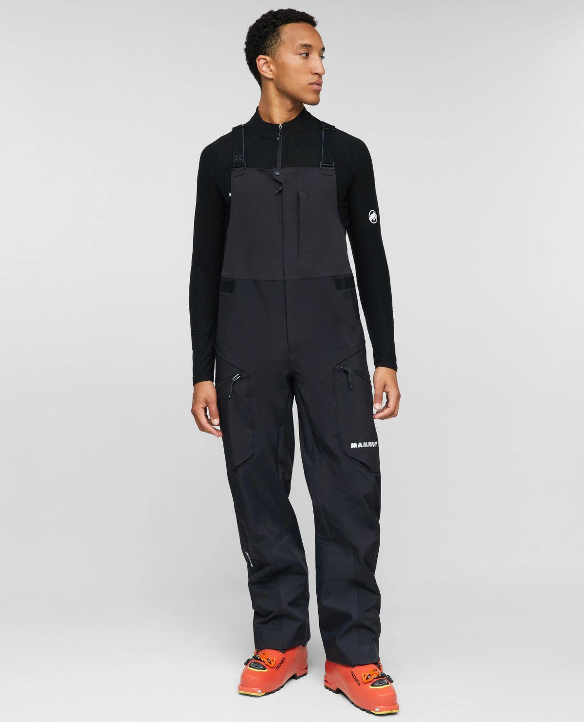 En modell i svarta Mammut Eiger Free Pro HS bib-skidbyxor och matchande överdel samt orange pjäxor.