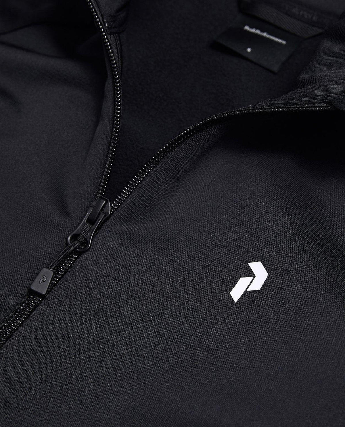 Närbild på dragkedja och bröstlogotyp på svart Peak Performance Rider Essentials Zip Jacket för dam.