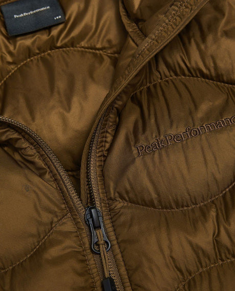 Närbild på Peak Performance Helium Down Vest Junior i brun färg med broderad logo och dragkedja.