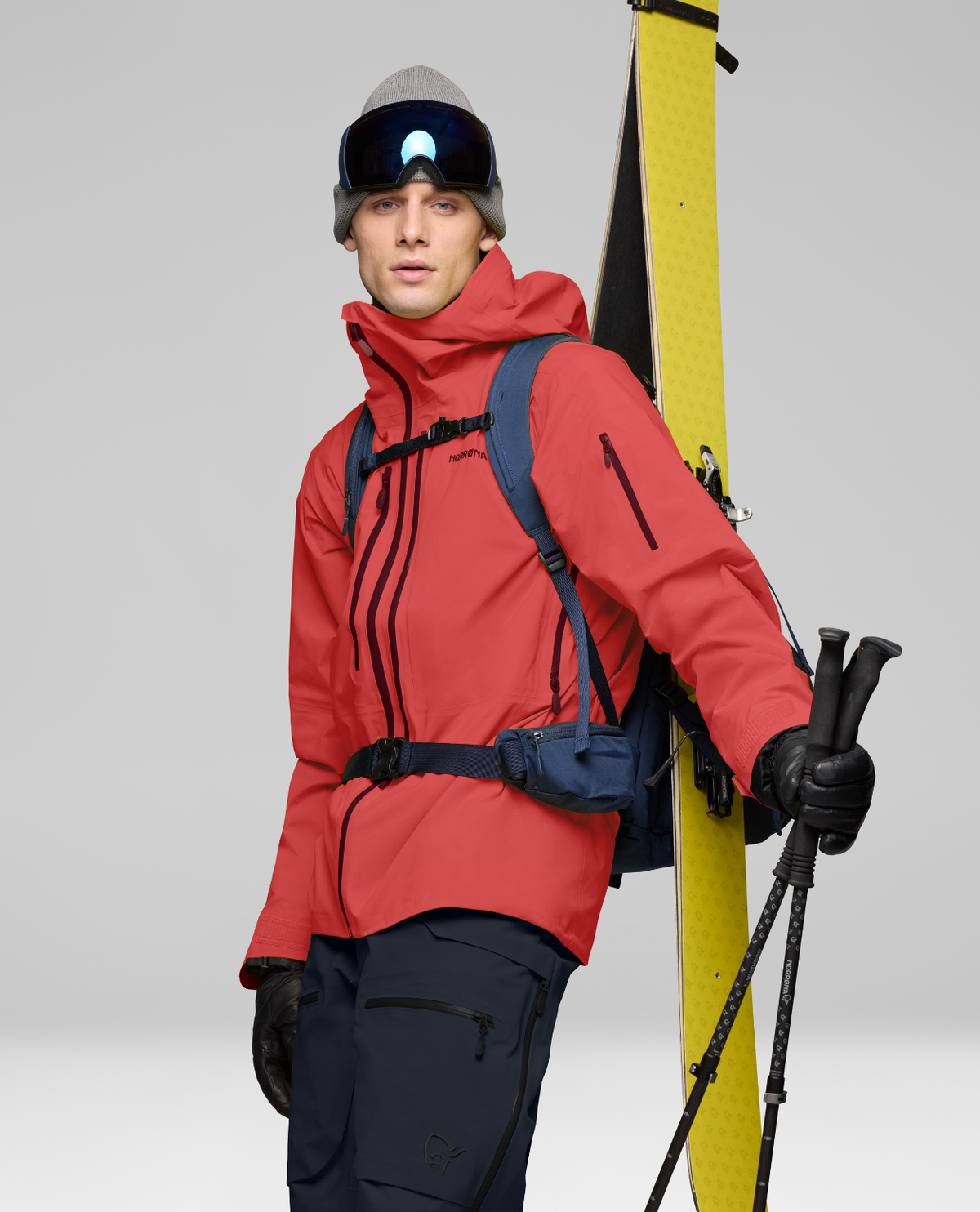 Norrøna Lofoten Gore-Tex Pro Jacket för herrar i den röda färgen Baked Apple, med skidåkare som håller skidor och stavar.