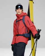 Norrøna Lofoten Gore-Tex Pro Jacket för herrar i den röda färgen Baked Apple, med skidåkare som håller skidor och stavar.
