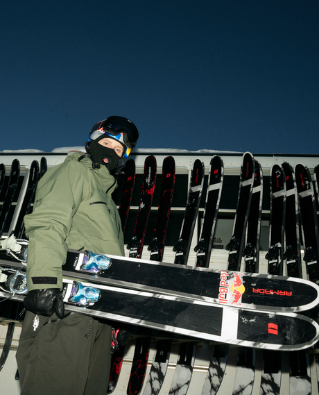Skidåkare i grön jacka och hjälm håller Armada ANTIMATTER 114 freerideskidor framför ett skidställ i snön.